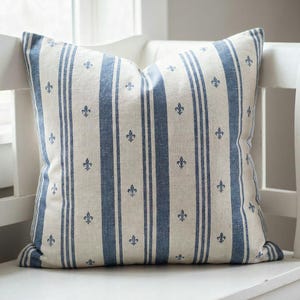 Close-up of the slate blue fleur de lis ticking stripe pillow cover on a white entryway bench