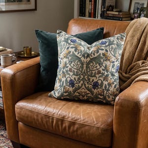 A botanical print throw pillow is centered on a brown leather armchair, accompanied by a dark green pillow and a tan knit throw blanket.