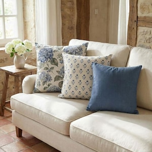 May include: Three square throw pillows arranged on a light beige sofa. One pillow features a blue hydrangea floral print, another has a small blue botanical pattern on a cream background, and the third is a solid blue textured fabric.