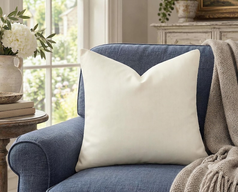 Solid warm ivory pillow cover on a slate blue armchair beside a window