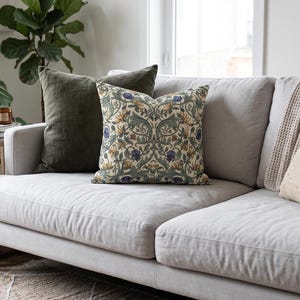 A light gray sectional sofa holds multiple decorative pillows including a botanical pattern square pillow, green velvet pillow, and mustard yellow cushion.