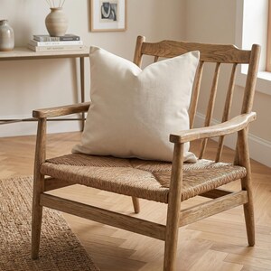 A wooden armchair with a woven seat and an ivory throw pillow on it, positioned in a well-lit room.