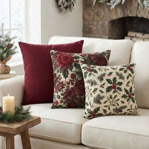 May include: Three square throw pillows arranged on a sofa. One pillow is solid deep burgundy. The second pillow features a dark floral pattern with red roses and greenery. The third pillow has a cream background with a holly and pine print.