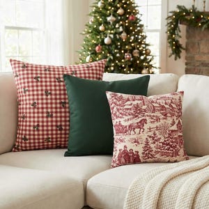 May include: Three square throw pillows arranged on a sofa. One pillow features a red and white gingham pattern with small holly sprigs, one is solid dark green, and one displays a red and white winter scene with figures and trees.