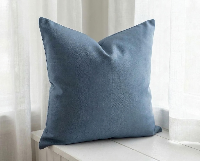 Close-up of the solid slate blue pillow cover on a window seat with sheer curtains, soft printed linen-look finish