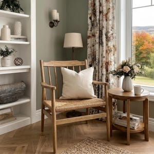 A wooden armchair with a woven seat and a solid ivory cushion, set in a cozy room with bookshelves and a window.