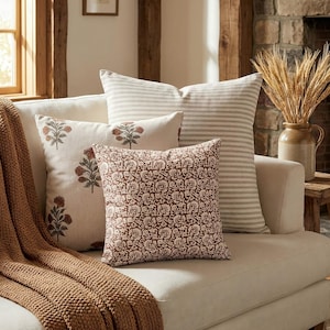 May include: A collection of three square throw pillows on a cream sofa. One pillow features thin horizontal beige stripes, one has a repeating floral block print in rust, and the front pillow displays a dense, intricate brown floral pattern.