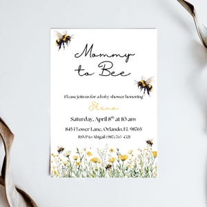 May include: A white rectangular baby shower invitation card featuring watercolour illustrations of bumblebees and wildflowers. The text reads Mommy to Bee, Please join us for a baby shower honouring Elena, Saturday, April 8th at 10 am, 845 Flower Lane, Orlando, FL 98765, RSVP to Abigail (987) 765-4321.