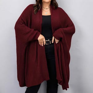 May include: A deep burgundy open-front cardigan featuring a chunky knit texture, long sleeves, and a relaxed, flowing silhouette. The garment is worn over a black top and black pants with a black belt featuring a silver-toned metal buckle.