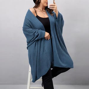 May include: A blue open-front knit cardigan with long sleeves and a textured pattern, worn over a black camisole and black trousers. The cardigan features a loose, relaxed fit with ribbed cuffs and a mid-thigh length, providing a layered look.