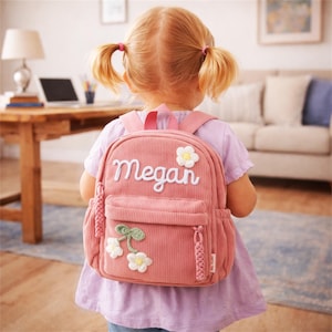 May include: A dusty pink corduroy backpack featuring the name Megan embroidered in white script. The bag includes a front zippered pocket decorated with white flower appliqués with yellow centers and green stems, plus braided zipper pulls.
