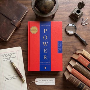 Könnte beinhalten: Ein gebundenes Exemplar von The 48 Laws of Power von Robert Greene mit rotem Einband und blauem Mittelstreifen, umgeben von einem Lederjournal, einem Globus, einem Tintenfass, einer Lupe, einem Füllfederhalter, einem Notizblock und alten Büchern.