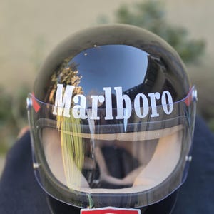 Può includere: Un casco da moto nero lucido con visiera trasparente e scritta Marlboro bianca sulla parte anteriore. Un logo rettangolare Texaco con bordi rossi e bianchi è posizionato sotto la visiera. L'imbottitura interna è di colore beige chiaro.