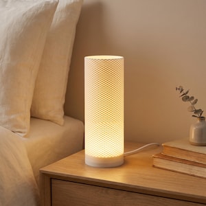 May include: A cylindrical table lamp with a white, textured, grid-patterned shade that emits a warm, soft glow. The lamp features a solid base and a white power cord extending from the back, providing illumination through its intricate surface.