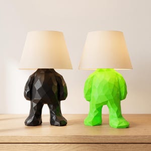 May include: Two table lamps featuring bases shaped like stylized, geometric figures with faceted surfaces. One base is matte black and the other is bright neon green. Each lamp is topped with a simple, tapered, off-white fabric lampshade.