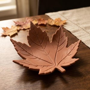 May include: A terracotta-colored bowl shaped like a maple leaf with detailed, recessed veins etched into the surface. The bowl features a pointed, multi-lobed edge and a small stem at the base, providing a shallow, open container for small items.