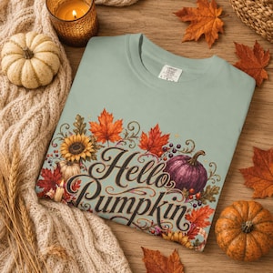 Puede incluir: Una camiseta de color verde salvia claro con un estampado gráfico que incluye el texto Hello Pumpkin en una fuente cursiva de color marrón oscuro. El diseño presenta ilustraciones de una calabaza morada, girasoles, hojas de arce y bayas.