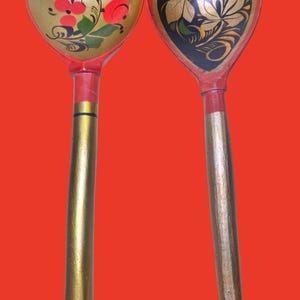 May include: Two wooden spoons with long handles. The left spoon features a gold bowl with red floral patterns and green leaves. The right spoon has a black bowl with gold and white floral designs. Both handles are painted in gold, red, and black.