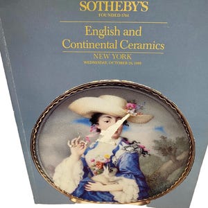 Può includere: Un catalogo d'asta Sotheby's intitolato English and Continental Ceramics, New York, Wednesday, October 26, 1988. La copertina presenta un'immagine circolare di una persona con un cappello di paglia e un abito blu che tiene un coniglio.