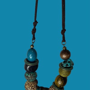 May include: A necklace featuring a brown cord with knots and a variety of beads in shades of turquoise, black, brown, and olive green. The beads include smooth, textured, and patterned spherical and oval shapes, some separated by small metallic rings.