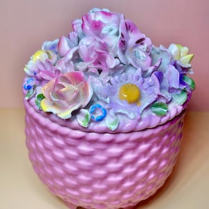 Puede incluir: Un joyero redondo de cerámica rosa con un patrón de tejido de cesta texturizado en la base. La tapa está adornada con un denso grupo de flores de porcelana esculpidas y multicolores en tonos morado, amarillo, azul y rosa con hojas verdes.