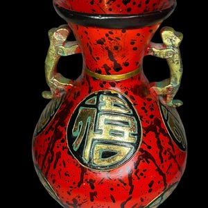 Può includere: Un vaso rosso con schizzi di vernice nera, dotato di due manici dorati a forma di creature stilizzate. La parte anteriore presenta un medaglione circolare nero contenente un grande carattere cinese dorato e dettagli dorati.