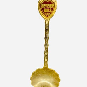 May include: A gold-toned metal souvenir spoon featuring a bamboo-style handle and a scalloped bowl. The top of the handle displays a shield-shaped emblem with a deep red background and the gold embossed text BEVERLY HILLS centered on the front.