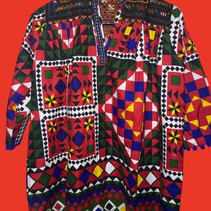 Puede incluir: Una túnica de manga larga con un vibrante patrón geométrico en rojo, verde, azul, amarillo y blanco. La prenda tiene un cuello mao con detalles bordados oscuros, un escote abierto y un dobladillo rematado con pequeños pompones azules.
