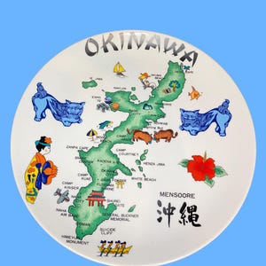 May include: A round white decorative plate featuring a green map of Okinawa with the text OKINAWA at the top and MENSOORE with Japanese characters below. The design includes blue shisa lion figures, a person in traditional clothing, and icons.