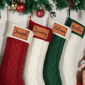 Peut inclure: Quatre chaussettes de Noël en tricot torsadé rouge, blanc et vert. Chaque chaussette comporte une étiquette en cuir beige personnalisée avec les prénoms Joseph, Quincy, Selina et Bella gravés en noir, fixée par des rivets métalliques.