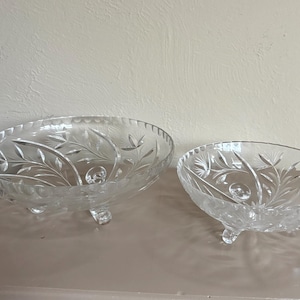 May include: Two clear glass bowls with three small feet each. Both bowls feature a cut glass pattern of leaves and stems. The larger bowl is oval-shaped with a scalloped rim, while the smaller bowl is round with a similar scalloped edge.
