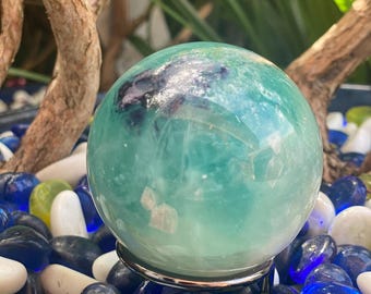 May include: A green and blue fluorite crystal sphere on a metal stand. The sphere is polished and has a swirling pattern of colors. The sphere is surrounded by small blue, green, and white stones.