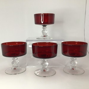 May include: A set of four matching dessert cups featuring deep red glass bowls atop clear glass stems and circular bases. Each cup has a wide, cylindrical bowl shape with a smooth rim, supported by a decorative, multi-tiered clear glass pedestal.