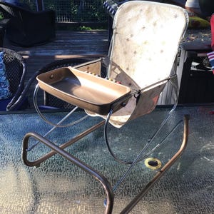 May include: A vintage-style rocking chair featuring a metallic bronze frame and a matching bronze tray. The seat and backrest are made of a light-colored fabric decorated with a repeating pattern of small, dark starburst shapes.