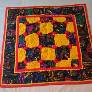 May include: A square quilt featuring a central grid of nine puffed yellow fabric squares separated by red fabric knots. The quilt is bordered by a vibrant, multi-coloured fabric with abstract patterns in shades of blue, purple, and green, finished with a red trim.