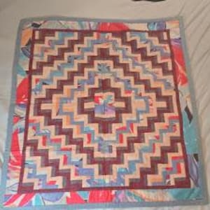 May include: A square quilt featuring a central geometric diamond pattern composed of alternating dark maroon and light beige fabric strips. The border displays a vibrant floral print with shades of red, blue, and teal, finished with a light blue binding.