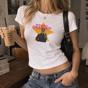 Może przedstawiać: Biały t-shirt z krótkim rękawem z żółtą grafiką gwiazdy z napisem "WHAT WOULD CHUCK DO?" oraz czarno-białym zdjęciem osoby w okularach przeciwsłonecznych, zestawiony z niebieskimi dżinsami i czarną torebką na ramię.
