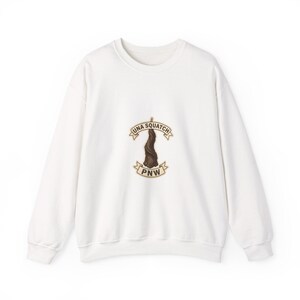 May include: A white long-sleeve crewneck sweatshirt featuring a central graphic of a brown, hairy, upright figure resembling a sasquatch. Above and below the figure are tan banners with the text UNA SQUATCH and PNW in dark brown lettering.
