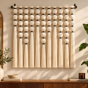 Puede incluir: Un tapiz de pared con diez hilos verticales suspendidos de una barra de madera horizontal. Cada hilo consta de una serie de cuentas esféricas de color blanco roto separadas por pequeños espaciadores negros, terminando en largas borlas.