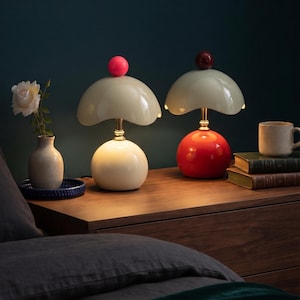 May include: Two table lamps with mushroom-shaped shades and spherical bases. The left lamp has a cream base and a bright pink sphere on top. The right lamp has a red base and a dark maroon sphere on top. Both feature gold-toned stems and light-colored shades.