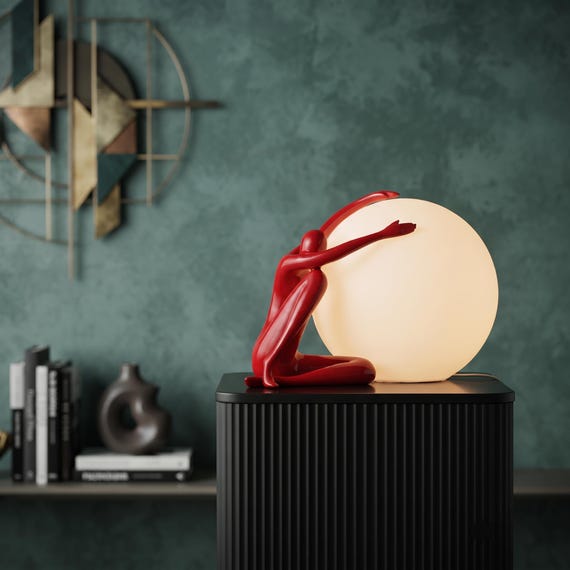 A minimalist human figure cradles a moon — sculptural resin table lamp that reads more like gallery art than a light fixture.