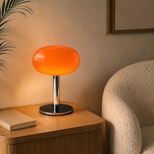 May include: A table lamp featuring a vibrant orange, rounded glass shade that emits a warm glow. The lamp has a polished chrome cylindrical stem and a circular black base, providing a sleek and balanced structure for this lighting fixture.