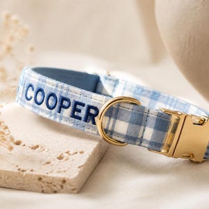 Op de afbeelding: Een huisdierhalsband met een lichtblauw en wit gingham-patroon, voorzien van de naam COOPER geborduurd in donkerblauw garen. De halsband heeft een gepolijste goudkleurige metalen gesp en een bijpassende goudkleurige D-ring voor de riem.