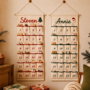 Könnte beinhalten: Zwei cremefarbene Wand-Adventskalender aus Stoff mit jeweils vierundzwanzig Taschen. Der linke Kalender zeigt den Namen Steven in Rot mit einem Baum-Symbol, der rechte Annie in Grün mit einem Weihnachtsmann-Symbol. Die Zahlen 1-24 sind aufgedruckt.