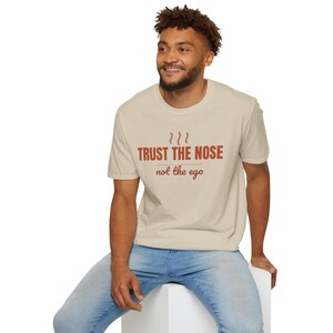 May include: A beige short-sleeved t-shirt featuring the text TRUST THE NOSE in bold brown capital letters above three wavy lines, with the phrase not the ego written in a smaller brown script font underneath, all centered on the chest area.