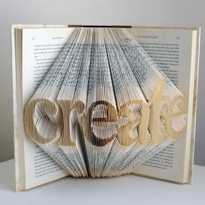 K&ouml;nnte beinhalten: Ein Buch mit dem Wort "create" in die Seiten gefaltet. Das Buch ist ge&ouml;ffnet und die Seiten sind gefaltet, um das Wort in 3D zu erstellen.
