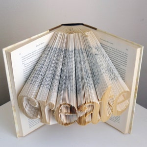 May include: An open book with pages folded to spell the word "CREATE". The book's pages are beige, and the text is black. The word "CREATE" is cut out of the pages, creating a 3D effect. The book is on a white surface.