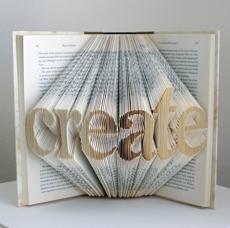 May include: An open book with the word "create" cut out of the pages. The pages are folded to create a diamond shape around the word. The book is open on a white surface.