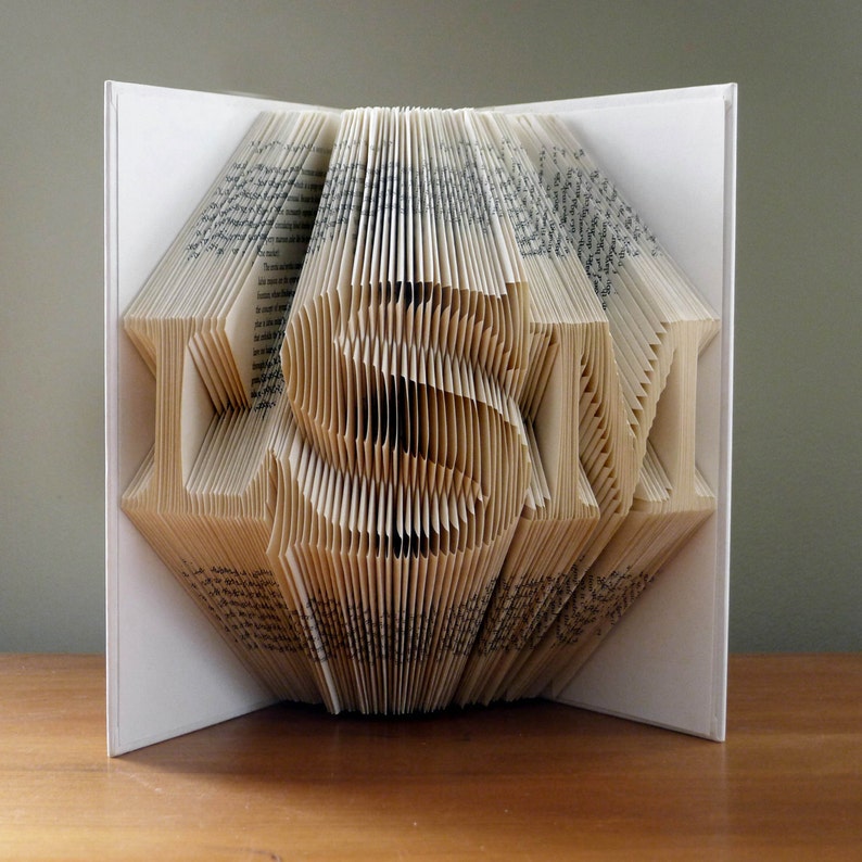 May include: A book folded into the shape of the word "LOVE" in a white cover. The book is made of pages with a cream colored background and black text.