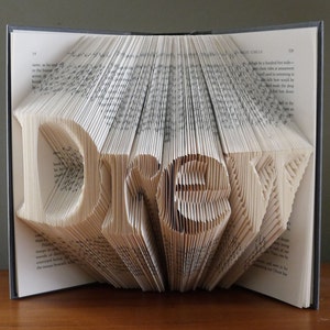 K&ouml;nnte beinhalten: Eine Buchkunstskulptur des Wortes "Dream", die durch das Falten der Seiten eines Buches entstanden ist. Das Buch ist ge&ouml;ffnet und die Seiten sind gefaltet, um die Buchstaben zu bilden. Das Buch liegt auf einer Holzoberfl&auml;che.